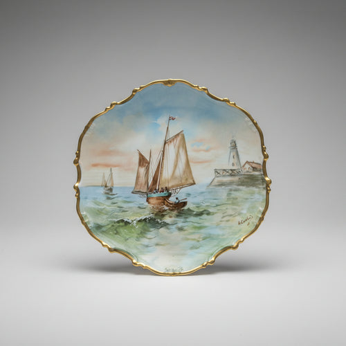 A scalloped ceramic plate with a gold-trimmed edge, featuring a hand painted nautical scene of sailboats on green-blue water under a pale sky.